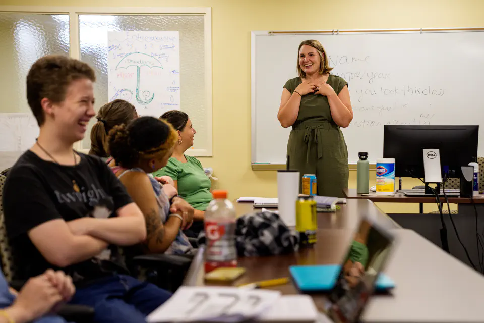 The image captures a lively classroom or workshop setting where a female instructor is enthusiastically engaging with her students, who appear amused and engaged.