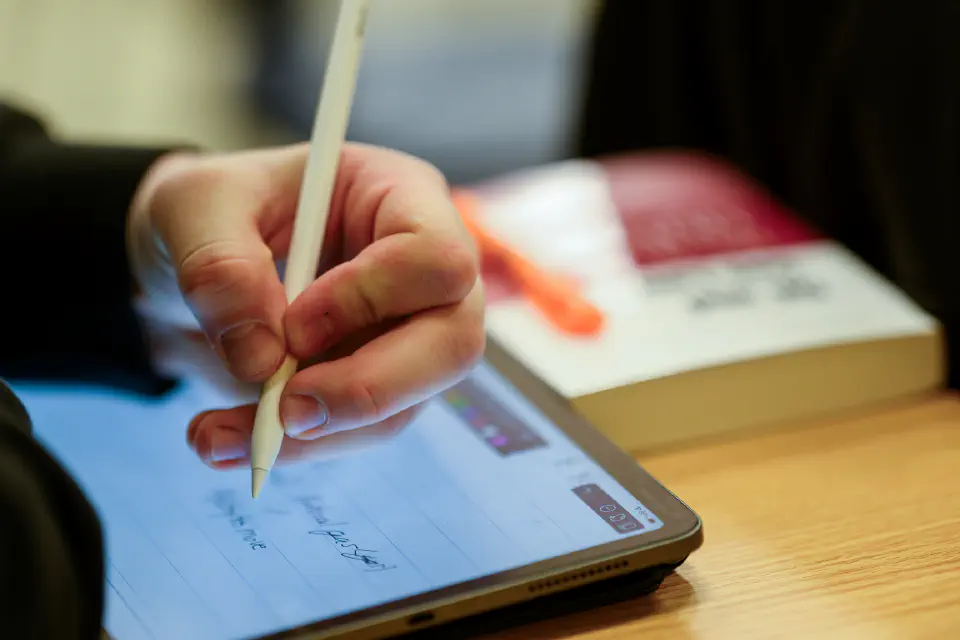 A person is using a stylus to write on a tablet, with a book in the background.